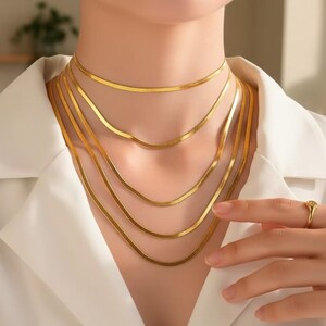 Op de afbeelding: Een verzameling goudkleurige visgraatkettingen van verschillende lengtes. De kettingen zijn gelaagd en worden gedragen met een wit overhemd met kraag. Een gouden ring is zichtbaar op de hand.