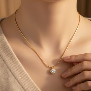 Peut inclure: Un collier doré avec un pendentif rond en pierre précieuse transparente. Le pendentif est serti dans une lunette dorée et suspendu à une fine chaîne dorée. Le collier est porté.