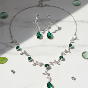 Può includere: Un set di collana e orecchini in argento con pietre preziose verde smeraldo a forma di goccia. La collana e gli orecchini presentano un delicato design a vite con piccoli cristalli trasparenti. Il set di gioielli è esposto su una superficie di marmo bianco.