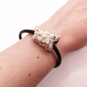 Può includere: Un bracciale con un cordino nero liscio e una perla centrale rettangolare in ceramica. La perla presenta una finitura smaltata color bianco sporco con crepe, macchie scure irregolari e venature distribuite sulla superficie ruvida.