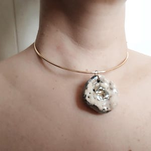 Può includere: Una collana girocollo in metallo color oro con un ciondolo singolo, irregolare e testurizzato. Il ciondolo presenta una superficie porosa color crema con un accento centrale color argento metallizzato, sospeso a una fascia sottile e lucida.