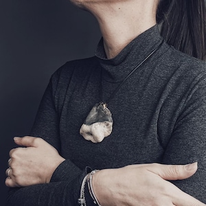 Puede incluir: Un collar con colgante único con una piedra grande de forma irregular. La piedra es una mezcla de gris y blanquecino, suspendida de un cordón fino y oscuro. El collar se lleva sobre un cuello alto gris oscuro.
