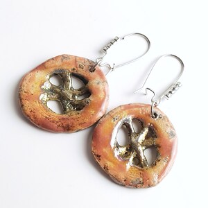 Può includere: Un paio di orecchini in ceramica fatti a mano. Ogni orecchino ha una forma circolare con un ritaglio a forma di croce al centro. Gli orecchini hanno una combinazione di colori rosso-arancio e oro con ganci per le orecchie argentati. Gli orecchini sono su sfondo bianco.