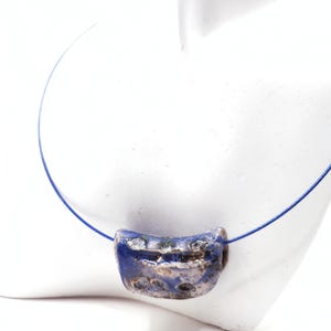 Può includere: Una collana con un ciondolo in ceramica rettangolare e ricurvo, caratterizzato da una finitura blu lucida e argento metallizzato, sospeso su un sottile filo blu flessibile. Il ciondolo presenta una superficie irregolare e testurizzata.