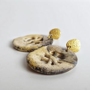 Puede incluir: Un par de pendientes de cerámica hechos a mano. Cada pendiente presenta un colgante circular blanquecino con un diseño tallado y un perno de color dorado. Los colgantes tienen un aspecto moteado y texturizado con acentos grises oscuros y marrones.