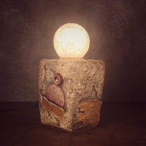 Könnte beinhalten: Eine Tischleuchte mit einem strukturierten, beigen, blockförmigen Sockel, der eingebettete kreisförmige und rechteckige Elemente in Rosa- und Goldtönen aufweist. Eine leuchtende, kugelförmige, durchscheinende weiße Glühbirne sitzt oben auf.