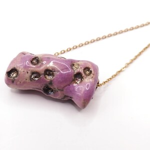 Può includere: Una collana con un ciondolo in ceramica cilindrico e irregolare, caratterizzato da uno smalto viola lavanda lucido con rientranze scure a forma di cratere sulla superficie, appeso a una sottile catenina in metallo dorato.