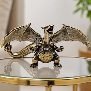 May include: A bronze dragon ornament with its wings spread and mouth open. The dragon is positioned on a round glass-topped table with a gold metal frame. The figurine has detailed scales and a long tail, and is a decorative item.