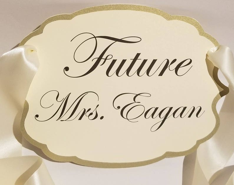 Puede incluir: Un cartel blanco con ribete dorado y una cinta de sat&eacute;n blanca. El cartel dice "Future Mrs. Eagan" en una fuente cursiva.