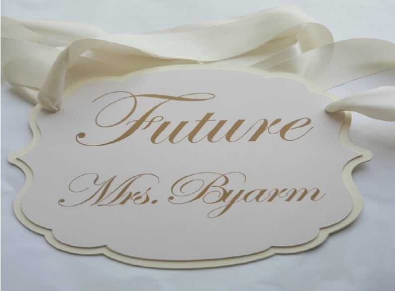 Puede incluir: Un cartel blanco con borde festoneado y una cinta dorada. El cartel dice "Future Mrs. Byars" en una fuente dorada cursiva.