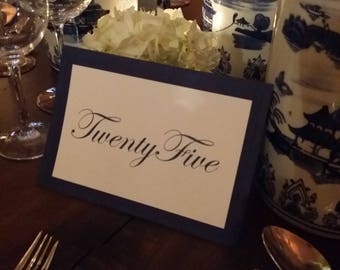 May include: A white card with a navy blue border and the text "Twenty-Five" in black cursive script. The card is sitting on a wooden table with a white and blue patterned plate and silverware.