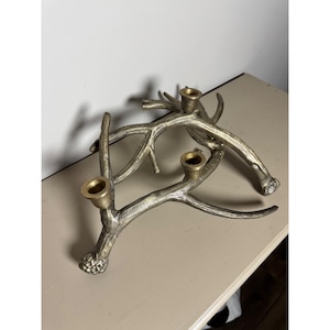 May include: A silver-toned metal candle holder in the shape of deer antlers. The candle holder has three brass-colored candle cups. The antlers are arranged in a naturalistic, branching pattern, creating a rustic and decorative accent piece.