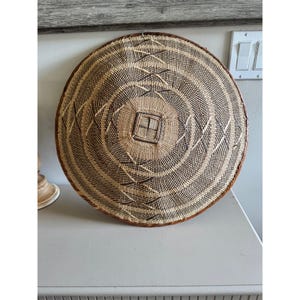 May include: A round woven basket with a brown rim and a square center design. The basket features concentric circles of natural and dark brown fibers. The design is symmetrical and appears to be handmade, suitable for home decor.