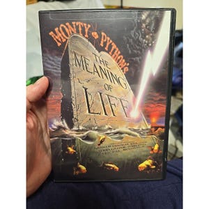 Pode incluir: Uma caixa de DVD de Monty Python's The Meaning of Life com um gráfico de uma tábua de pedra com texto, um raio e peixes a nadar na água. A capa inclui os nomes dos escritores: Graham Chapman, John Cleese, Terry Gilliam, Terry Jones, Eric Idle e Michael Palin.