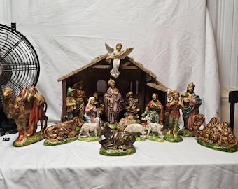 May include: A nativity scene set featuring a wooden stable with a straw roof and a central angel figure. The collection includes hand-painted figurines of Mary, Joseph, the infant Jesus, shepherds, wise men, camels, sheep, and a donkey on a white surface.