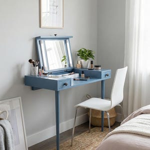 Puede incluir: Tocador azul con espejo abatible y dos cajones. El tocador tiene una silla blanca, una planta pequeña y artículos de maquillaje encima. Una foto enmarcada cuelga de la pared encima del tocador.