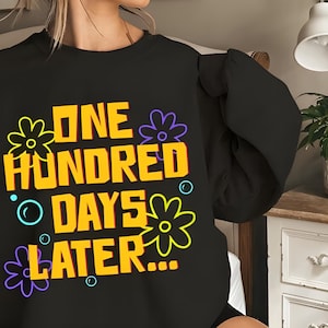 May include: Black sweatshirt with the text "ONE HUNDRED DAYS LATER..." in yellow, outlined in orange, with flower and bubble graphics. The sweatshirt is a casual style.