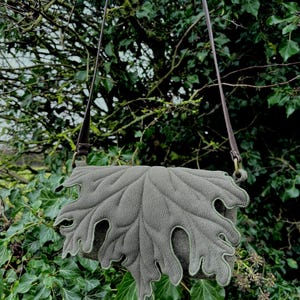 May include: A grey shoulder bag in the shape of a leaf, featuring detailed stitching to mimic leaf veins and a green trim. It has a long, brown strap. The bag is set against a backdrop of green foliage.