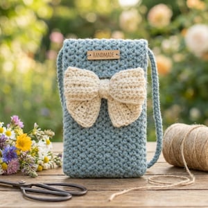 May include: A handmade, light blue crochet phone bag with a cream-colored bow. The bag has a long strap and a small wooden tag that reads "HANDMADE". The bag is sitting on a wooden surface with flowers and scissors in the background.