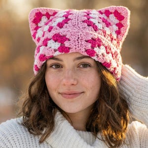 May include: A pink, white, and fuchsia crochet cat ear hat. The hat is made of granny squares and is worn by a person wearing a white sweater. The hat has two pointed ears on top.