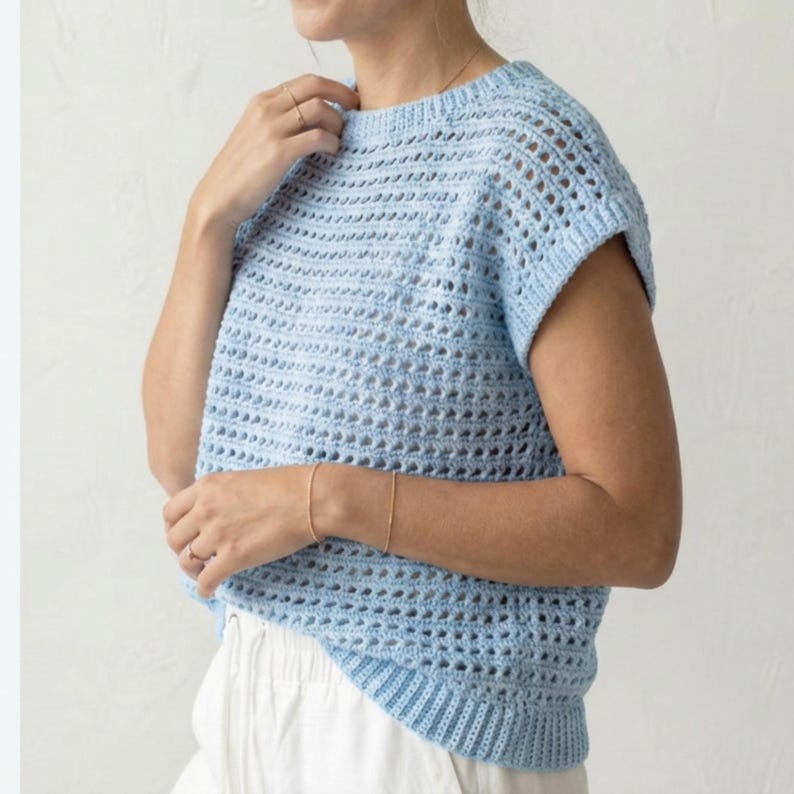Puede incluir: Un top de crochet azul claro con cuello redondo y mangas cortas. El dise&ntilde;o de tejido abierto crea un aspecto aireado y transpirable. El top es de color liso y parece hecho a mano. El dobladillo inferior y los pu&ntilde;os de las mangas tienen una textura acanalada.
