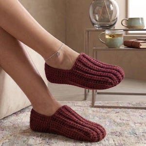 Puede incluir: Un par de zapatillas de punto color burdeos. Las zapatillas tienen una textura acanalada y un diseño de punta cerrada. La imagen muestra las zapatillas en los pies de una persona, lo que sugiere comodidad y calidez. Un accesorio acogedor para uso en interiores.