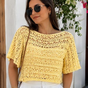 Puede incluir: Una blusa de ganchillo en color amarillo brillante con un amplio escote barco y mangas cortas acampanadas. La prenda está confeccionada con un intrincado patrón calado que alterna bandas horizontales de tejido delicado y filas sólidas.