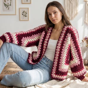 Può includere: Un cardigan all'uncinetto fatto a mano, senza chiusura, con un motivo a granny square. Il maglione presenta un design a blocchi di colore con sfumature di bordeaux, rosa e crema. Le maniche sono lunghe e voluminose. La persona indossa jeans blu.