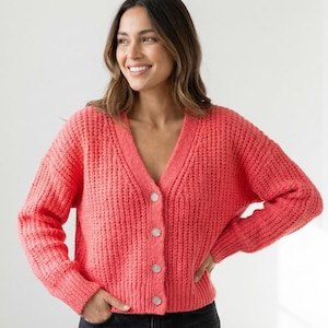 May include: A coral-colored, chunky knit cardigan with a V-neck and button closure. The sweater has long sleeves and a ribbed texture. The buttons are a light, neutral color. The cardigan is a casual, everyday garment.