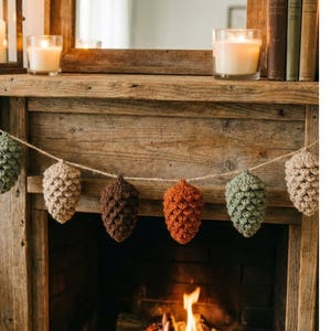 May include: A rustic fireplace mantel with a garland of crocheted pinecones in shades of green, brown, and beige. Lit candles in glass holders and a mirror are visible. The fireplace below has a burning fire.