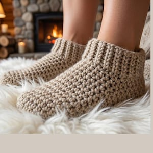 May include: A close-up shot of beige crochet slipper boots. The boots feature a ribbed cuff and a textured stitch. They are placed on a fluffy white surface, with a fireplace and candles visible in the background.