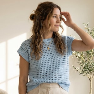 Puede incluir: Top de crochet azul claro con cuello redondo y mangas cortas. El top tiene un diseño de tejido abierto y texturizado. La persona lleva un collar y una pulsera dorados. El top es de color azul claro.