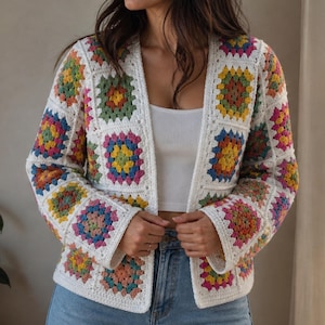Puede incluir: Un cárdigan de crochet blanco con patrones cuadrados coloridos. Los cuadrados presentan una variedad de colores, incluyendo amarillo, rosa, verde y azul. El cárdigan tiene mangas largas y un cuello.