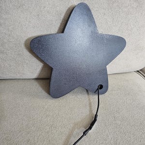 Op de afbeelding: Een donkergrijs, stervormig object met een textuuroppervlak. Een zwarte draad loopt uit een gat aan de onderkant van de ster. De ster is waarschijnlijk een decoratief item of onderdeel van een groter ontwerp.