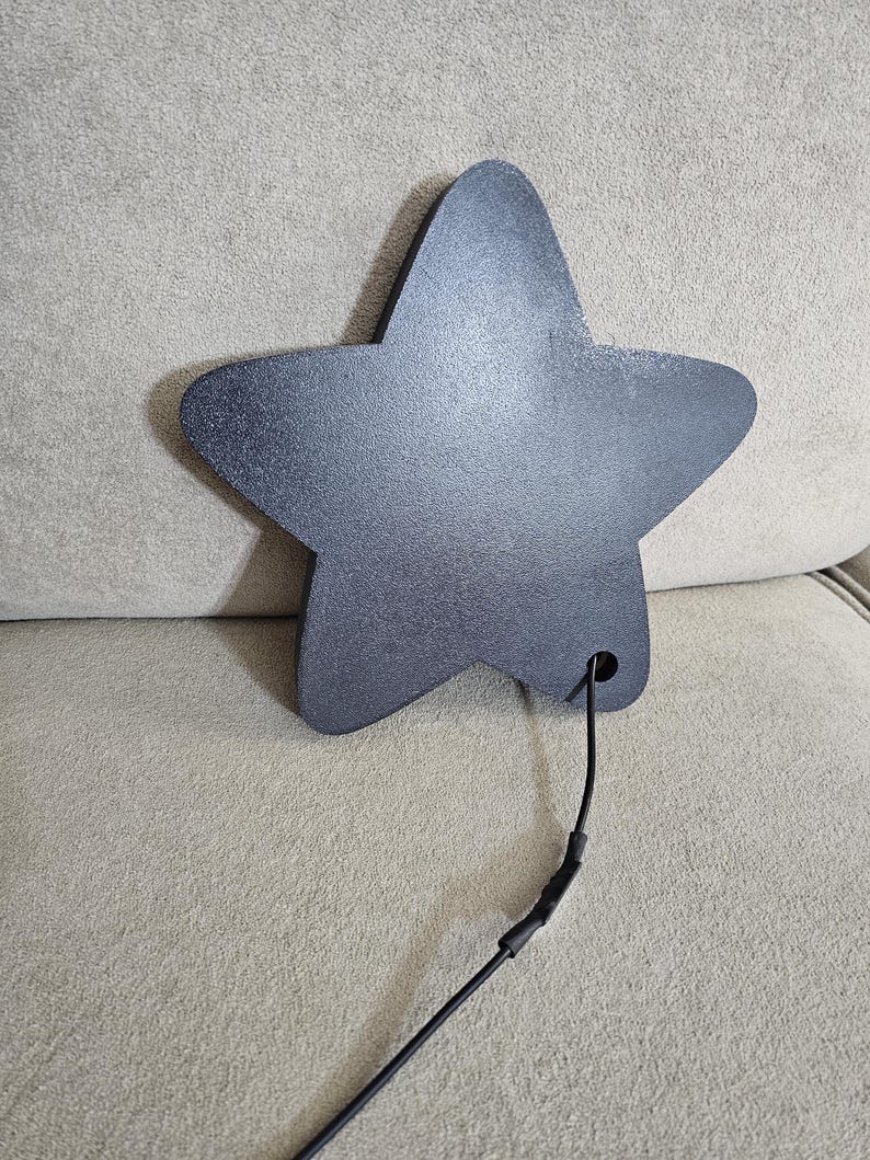 Op de afbeelding: Een donkergrijs, stervormig object met een textuuroppervlak. Een zwarte draad loopt uit een gat aan de onderkant van de ster. De ster is waarschijnlijk een decoratief item of onderdeel van een groter ontwerp.