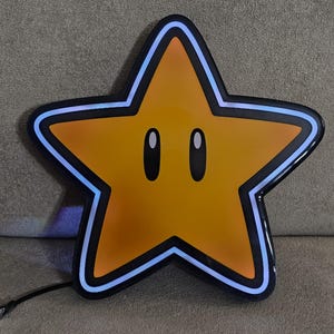 Op de afbeelding: Een grote, stervormige lamp met een gele kern en zwarte omtrek. De ster heeft twee ovale ogen en een blauwe neonrand. Een zwart snoer loopt vanaf de onderkant van de ster.