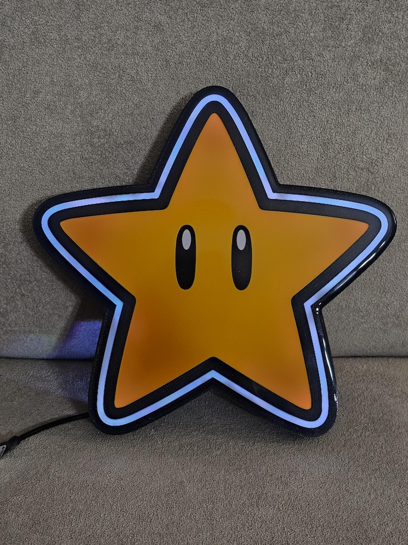 Op de afbeelding: Een grote, stervormige lamp met een gele kern en zwarte omtrek. De ster heeft twee ovale ogen en een blauwe neonrand. Een zwart snoer loopt vanaf de onderkant van de ster.