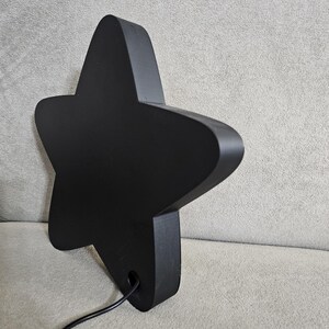 Op de afbeelding: Een zwarte, stervormige decoratieve lamp met afgeronde randen. De lamp heeft een matte afwerking en een netsnoer dat uit de basis komt. De ster staat schuin tegen een neutrale achtergrond.