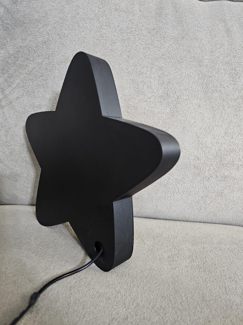 Op de afbeelding: Een zwarte, stervormige decoratieve lamp met afgeronde randen. De lamp heeft een matte afwerking en een netsnoer dat uit de basis komt. De ster staat schuin tegen een neutrale achtergrond.