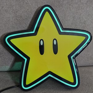 Op de afbeelding: Een geel stervormig licht met een zwarte rand en een lichtgroene omtrek. De ster heeft twee ovale ogen en een zwart netsnoer. Het licht is een decoratief item.