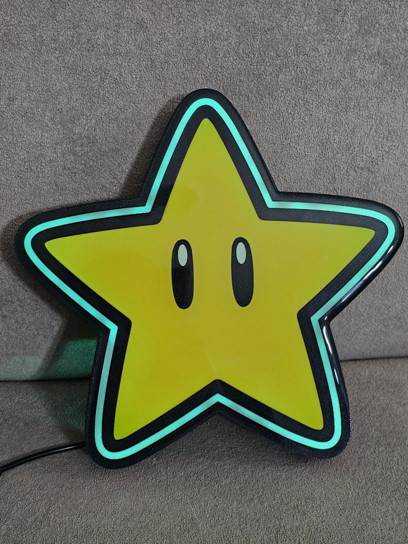 Op de afbeelding: Een geel stervormig licht met een zwarte rand en een lichtgroene omtrek. De ster heeft twee ovale ogen en een zwart netsnoer. Het licht is een decoratief item.