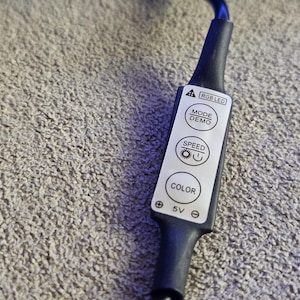 Op de afbeelding: Zwarte RGB LED-controller met witte knoppen met de tekst "MODE DEMO", "SPEED" en "COLOR". De controller is aangesloten op een zwarte kabel. De achtergrond is een grijs, getextureerd oppervlak.