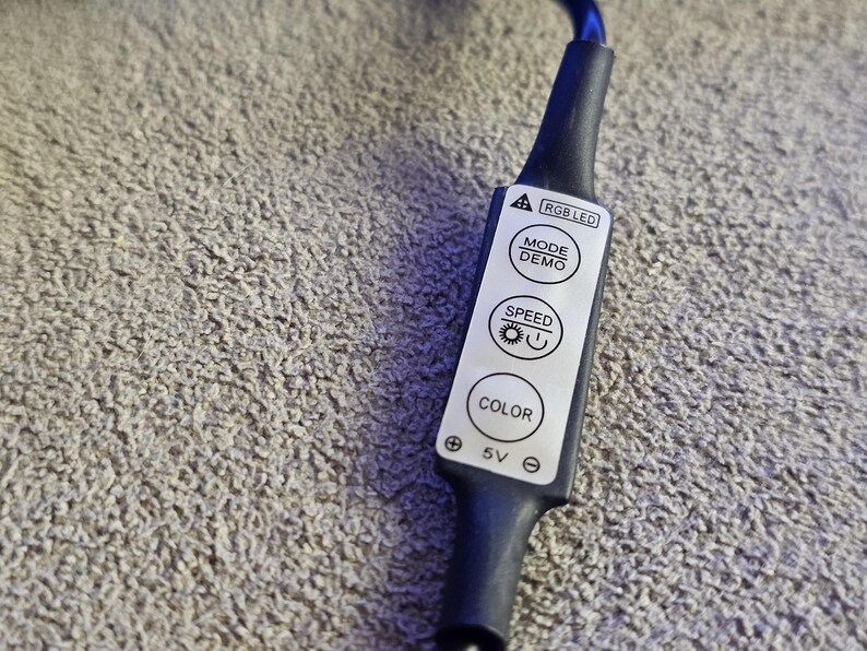 Op de afbeelding: Zwarte RGB LED-controller met witte knoppen met de tekst "MODE DEMO", "SPEED" en "COLOR". De controller is aangesloten op een zwarte kabel. De achtergrond is een grijs, getextureerd oppervlak.