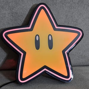 Op de afbeelding: Een grote, stervormige lamp met een cartoongezicht. De ster is voornamelijk oranje met een zwarte omtrek en roze rand. Hij heeft twee ovale ogen en een lachende uitdrukking. Een zwart netsnoer loopt van de onderkant.