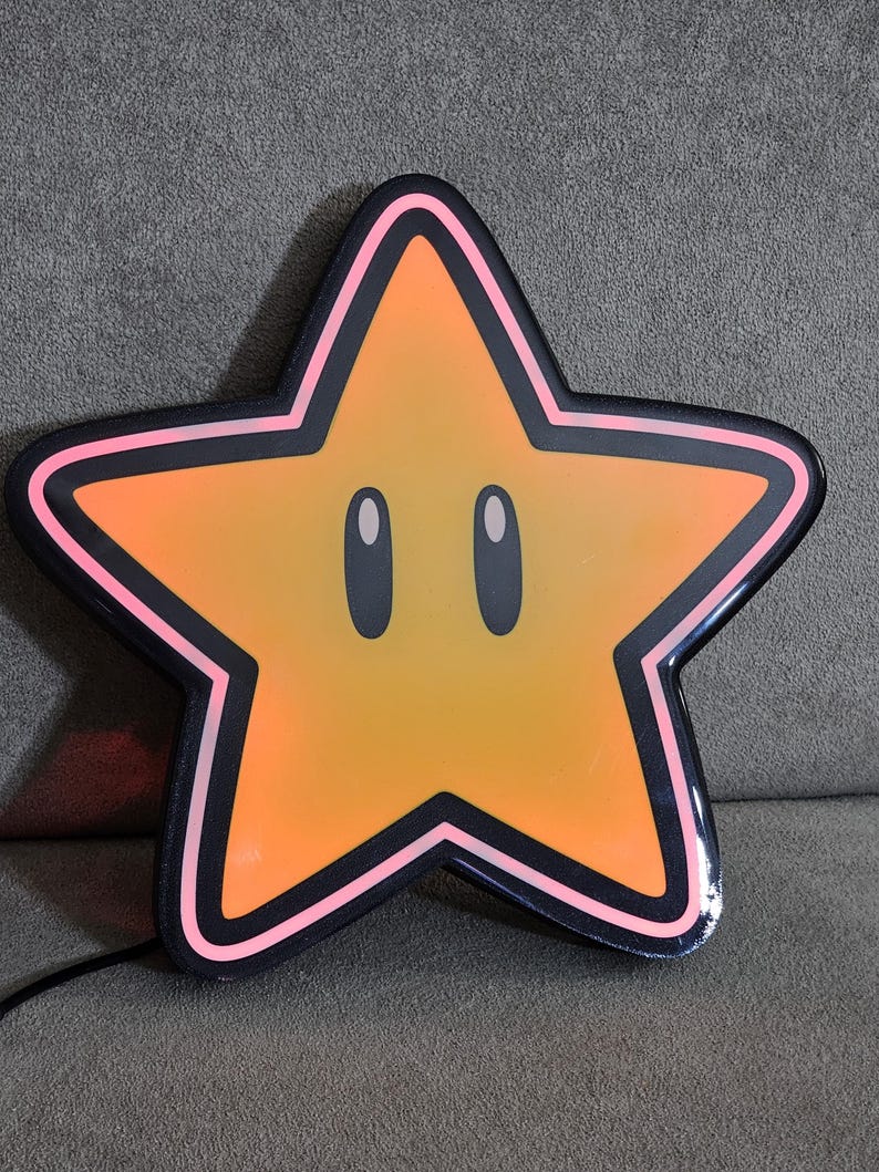 Op de afbeelding: Een grote, stervormige lamp met een cartoongezicht. De ster is voornamelijk oranje met een zwarte omtrek en roze rand. Hij heeft twee ovale ogen en een lachende uitdrukking. Een zwart netsnoer loopt van de onderkant.