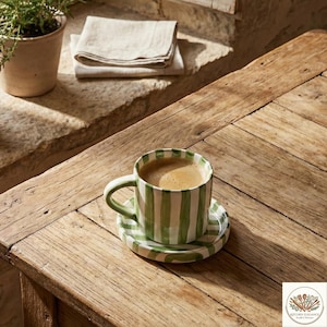 Puede incluir: Una taza y un platillo de caf&eacute; de cer&aacute;mica a rayas verdes y blancas llenas de caf&eacute;. La taza y el platillo est&aacute;n sobre una mesa de madera r&uacute;stica. Una peque&ntilde;a maceta y servilletas de lino dobladas est&aacute;n en el fondo.