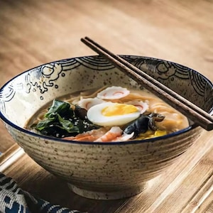 May include: A bowl of ramen with chopsticks. The bowl has a speckled beige exterior with a blue patterned rim. The ramen includes noodles, a boiled egg, shrimp, seaweed, and fish cakes. The chopsticks are brown.