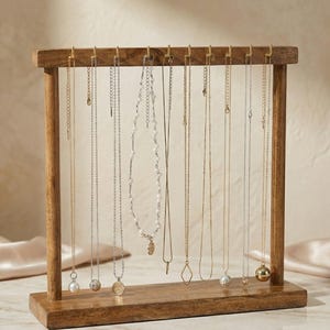 May include: A wooden jewelry display stand with multiple necklaces hanging from it. The necklaces are of varying lengths and styles, including chains, pearls, and pendants. The stand is made of light brown wood and has a rectangular base.