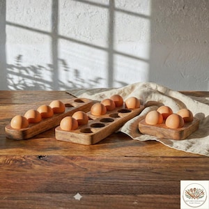 Könnte beinhalten: Eierhalter aus Holz mit braunen Eiern. Die Halter sind rechteckig mit geschnitzten, eiförmigen Vertiefungen. Die Eier sind hellbraun und liegen in den geschnitzten Aussparungen. Auf einem Holztisch mit weißem Tuch.