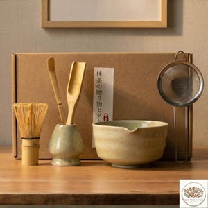 May include: A matcha tea set featuring a bamboo whisk, ceramic bowl, wooden scoop, and a metal sieve. The set includes a small ceramic vase holding two wooden utensils. The box behind the set has Japanese text.